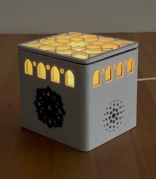 AdhanBox allumé sur une table en bois : les arches et le couvercle ajouré laissent passer une lumière chaude, un motif géométrique orne la façade.